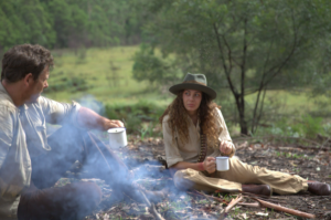 White man at campfire, adjecent, Jarrah (woman) drinks from a cup