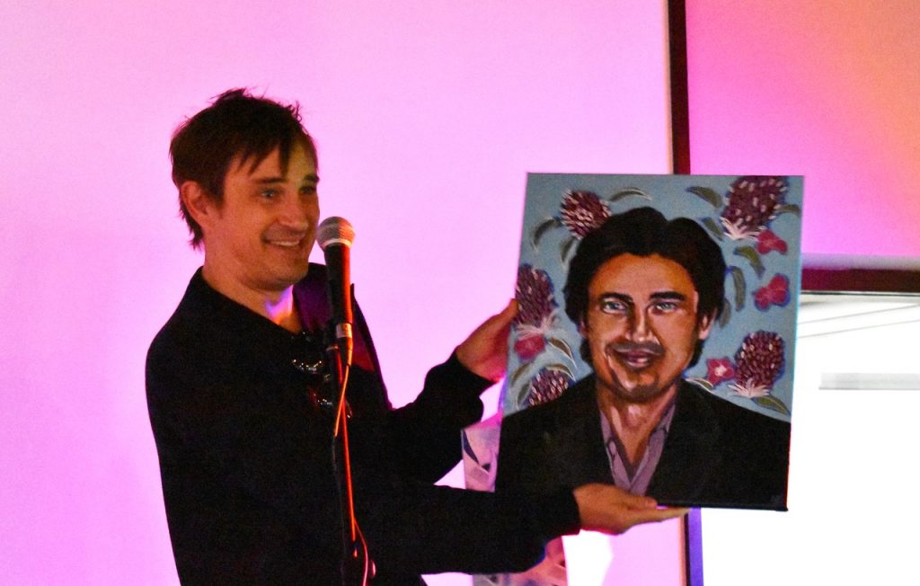 Magenta background. Trent Dalton (man) stands behind microphone on stage, holding a self portrait.