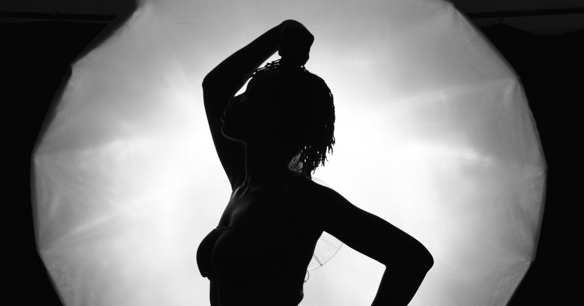 A black and white image of a woman standing in front of a large, white light. She is posing seductively, with one arm resting on her head and the other hand on her lower back.
