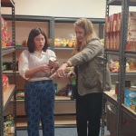 Two women standing in a storage room, packing away groceries. One woman directing the other.