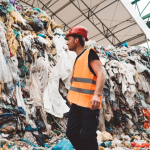 A worker wearing a hi-vis vest standing in front a pile of discarded clothing, rotting away in a landfill.