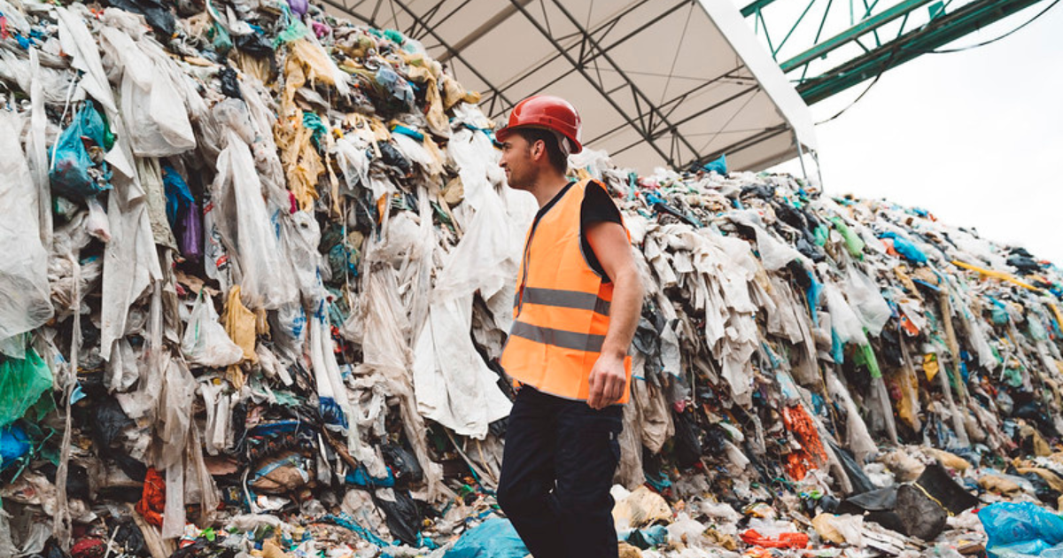 A worker wearing a hi-vis vest standing in front a pile of discarded clothing, rotting away in a landfill.
