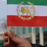 A photo of a hand holding a small Iranian flag that has a sun and lion depicted on it.